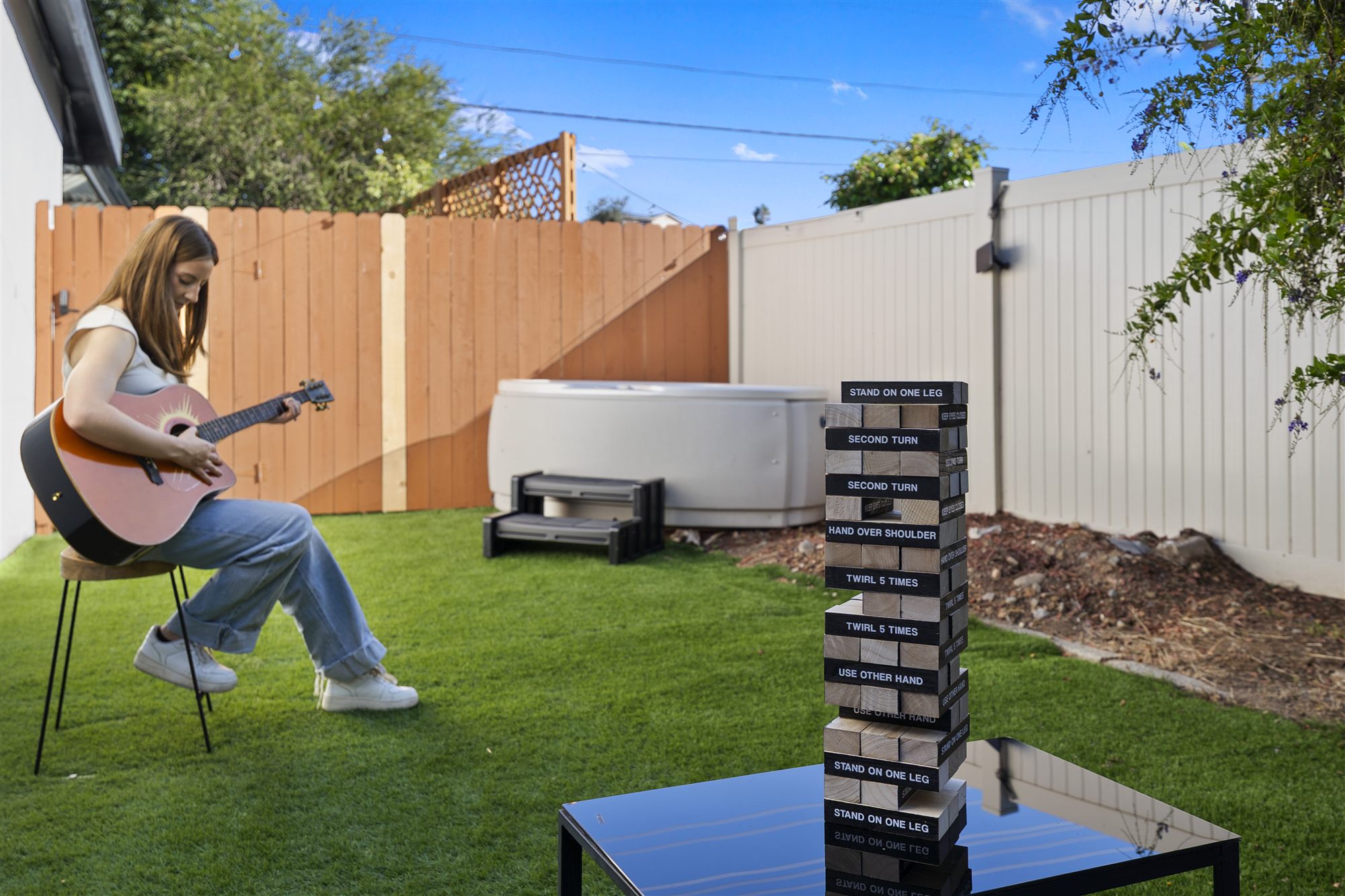 Woman playing guitar on the grass with giant Jenga mid-game and the hot tub behind